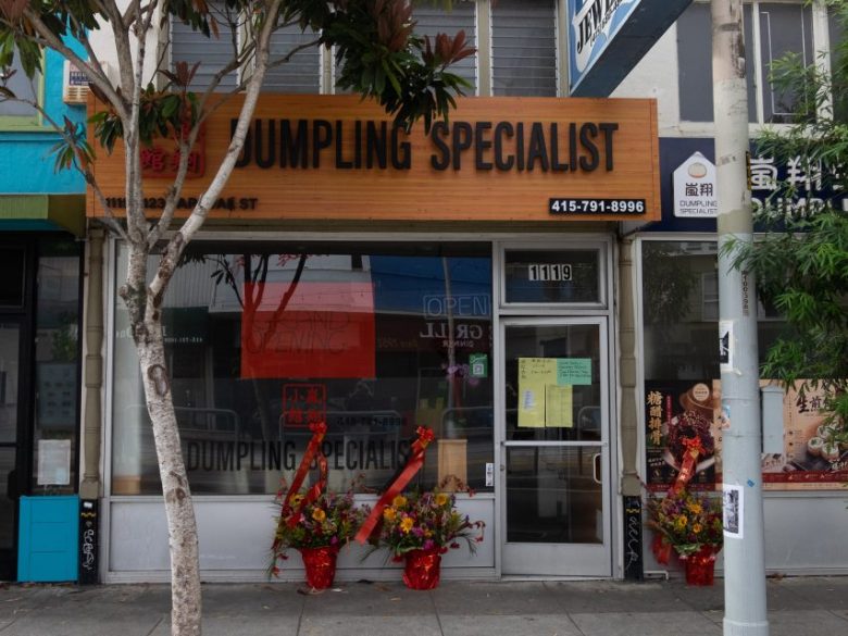 Street view of a closed dumpling restaurant with flower stands outside, a wooden sign reading “Dumpling Specialist,” and a yellow notice posted on the glass door.