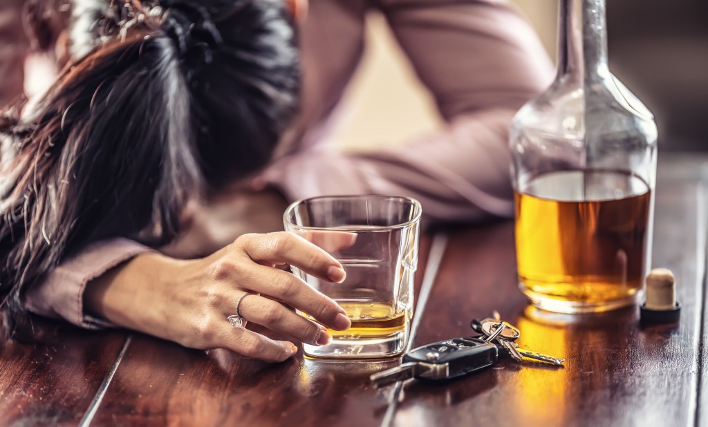 A woman in a pink shirt passed out on a table with a glass of whiskey, a bottle of whiskey, and car keys.