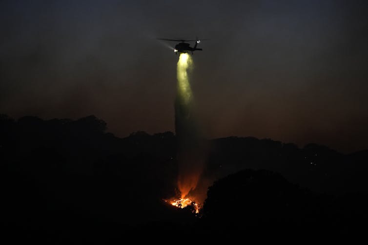 A fire helicopter drops water over flames in the dark as crews battle a wildfire.