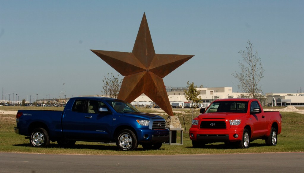 Two Toyota Tundra pickup trucks, one blue and one red, parked in front of a large metal star sculpture, with the assembly plant in the background.