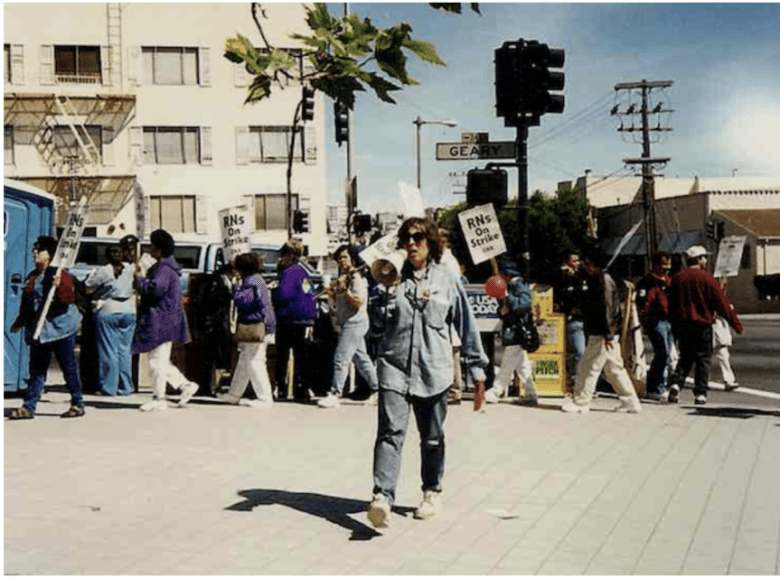 A person with a megaphone leads a group of people holding "RNs On Strike" signs on a city street corner near a building and traffic lights.