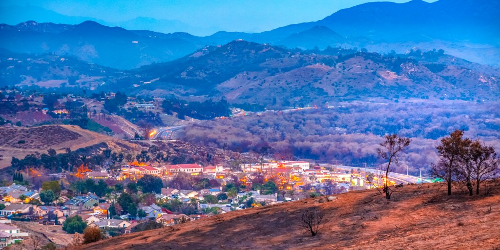 View of homes in Fallbrook, California, near hills destroyed by the Lilac Fire, with burnt ground and trees.