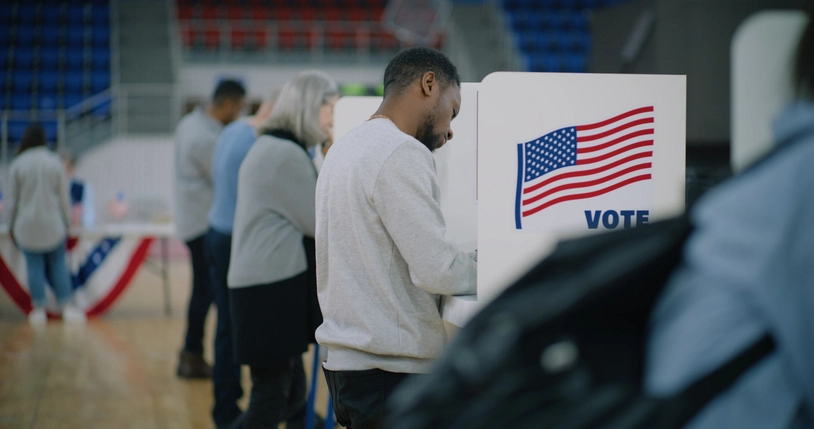 A voter casts a ballot at a polling place during an election, with voting booths and other voters visible in the background.
