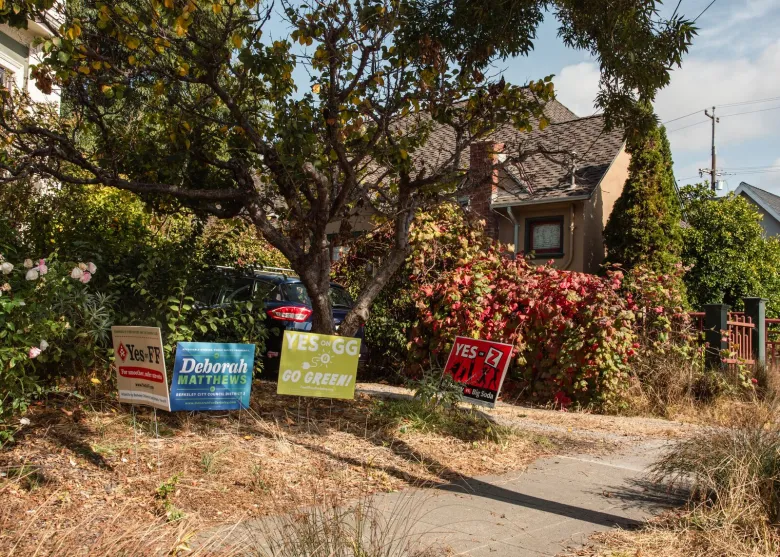 Four signs for various Berkeley campaigns are planted in the yellowed grass of a front yard.