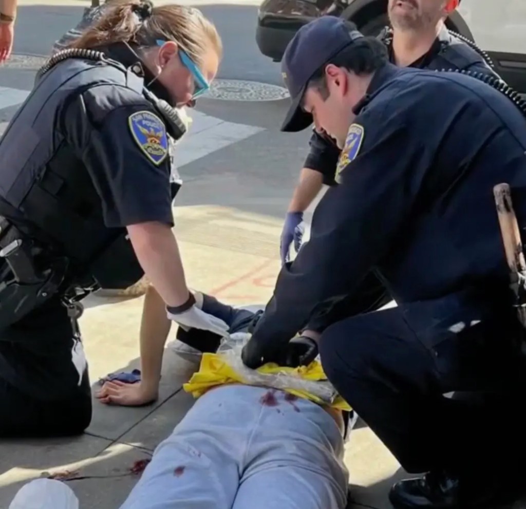 Police officers assisting a stabbing victim lying on the ground, with visible blood.