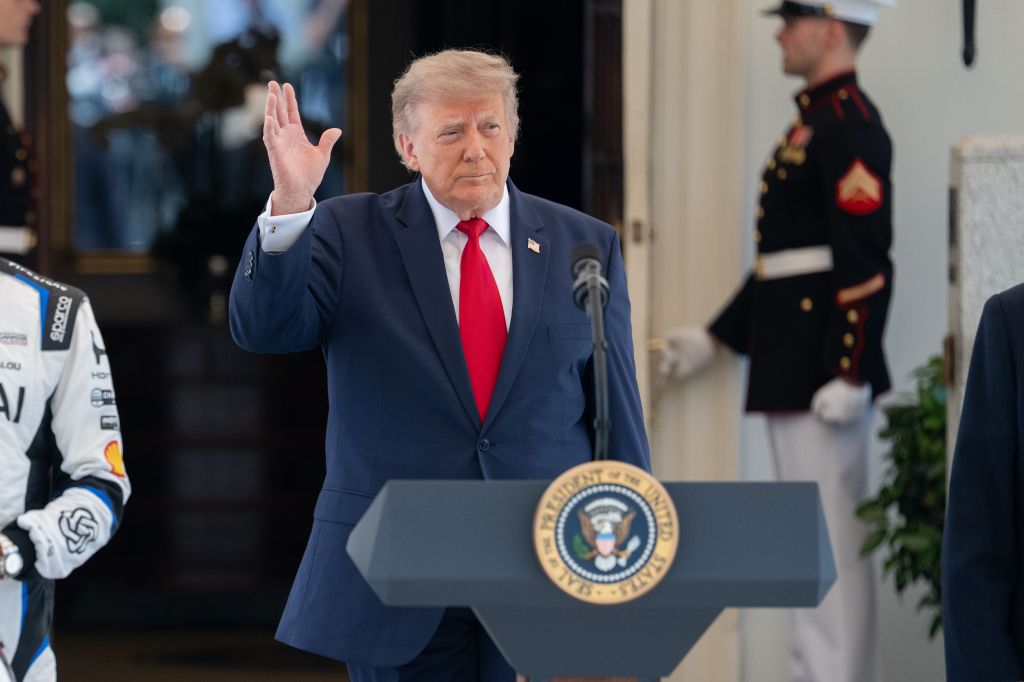 Donald Trump raises his hand while speaking at a podium with the Presidential Seal.