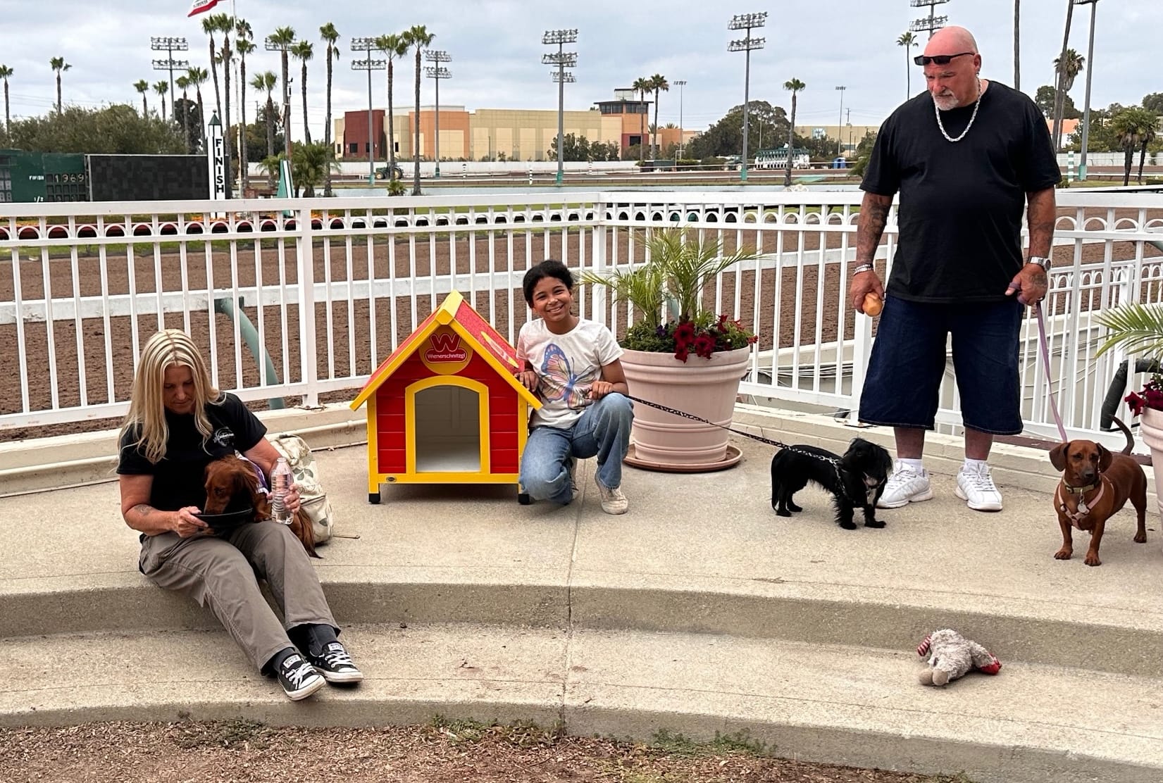 People sit and stand near a small red and yellow dog house.