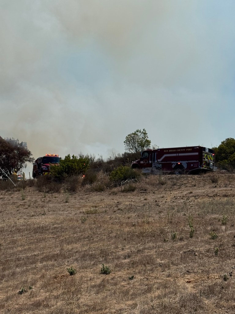Two fire trucks are parked in a field with dry grass, with a plume of smoke in the background.