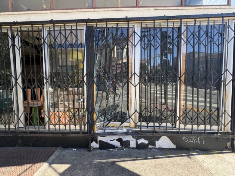 Storefront with metal security bars, a broken glass window, and damage to the lower wall. Reflections and street scene visible in the window. Graffiti is written on the bottom right corner.