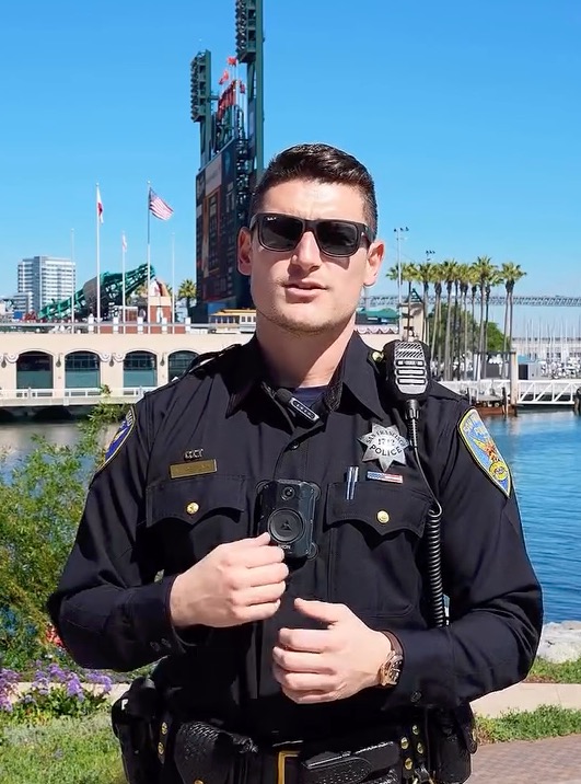 San Francisco Police Officer Mike Petuya in uniform with a body camera, in front of a stadium and body of water.