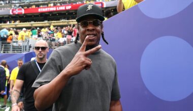 Jay-Z, wearing sunglasses and a cap, flashes a peace sign at an event, with a crowd in the background.