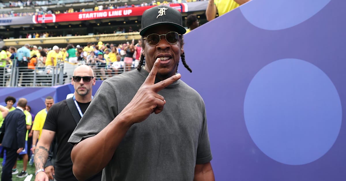 Jay-Z, wearing sunglasses and a cap, flashes a peace sign at an event, with a crowd in the background.