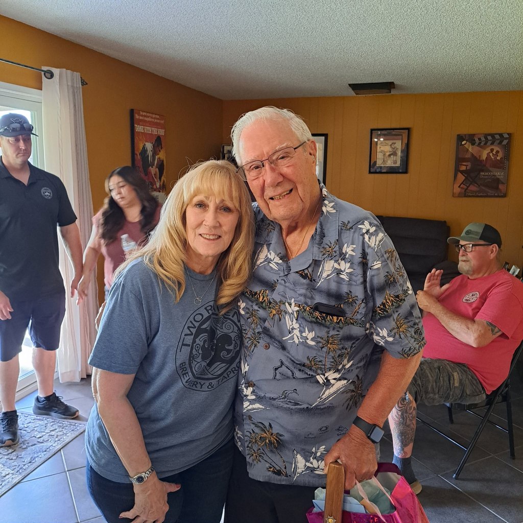 A blonde woman in a blue t-shirt with a gray-haired man in a Hawaiian shirt holding a wooden walking cane.