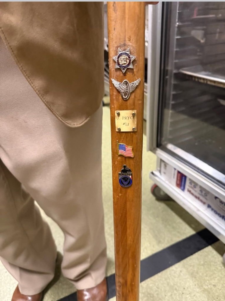 A walking cane with an array of military and police badges, a US flag pin, and a plaque with "J. Pryce #L1" inscribed on it.
