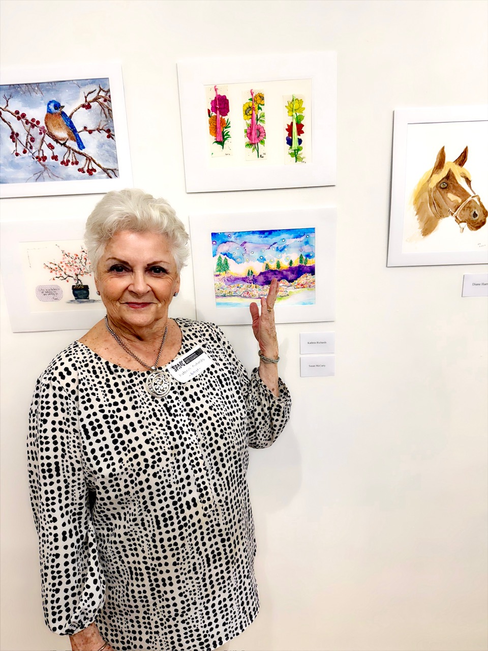 Older woman with short white hair in a black-and-white polka-dot blouse stands beside framed artworks in a gallery, smiling at the camera.