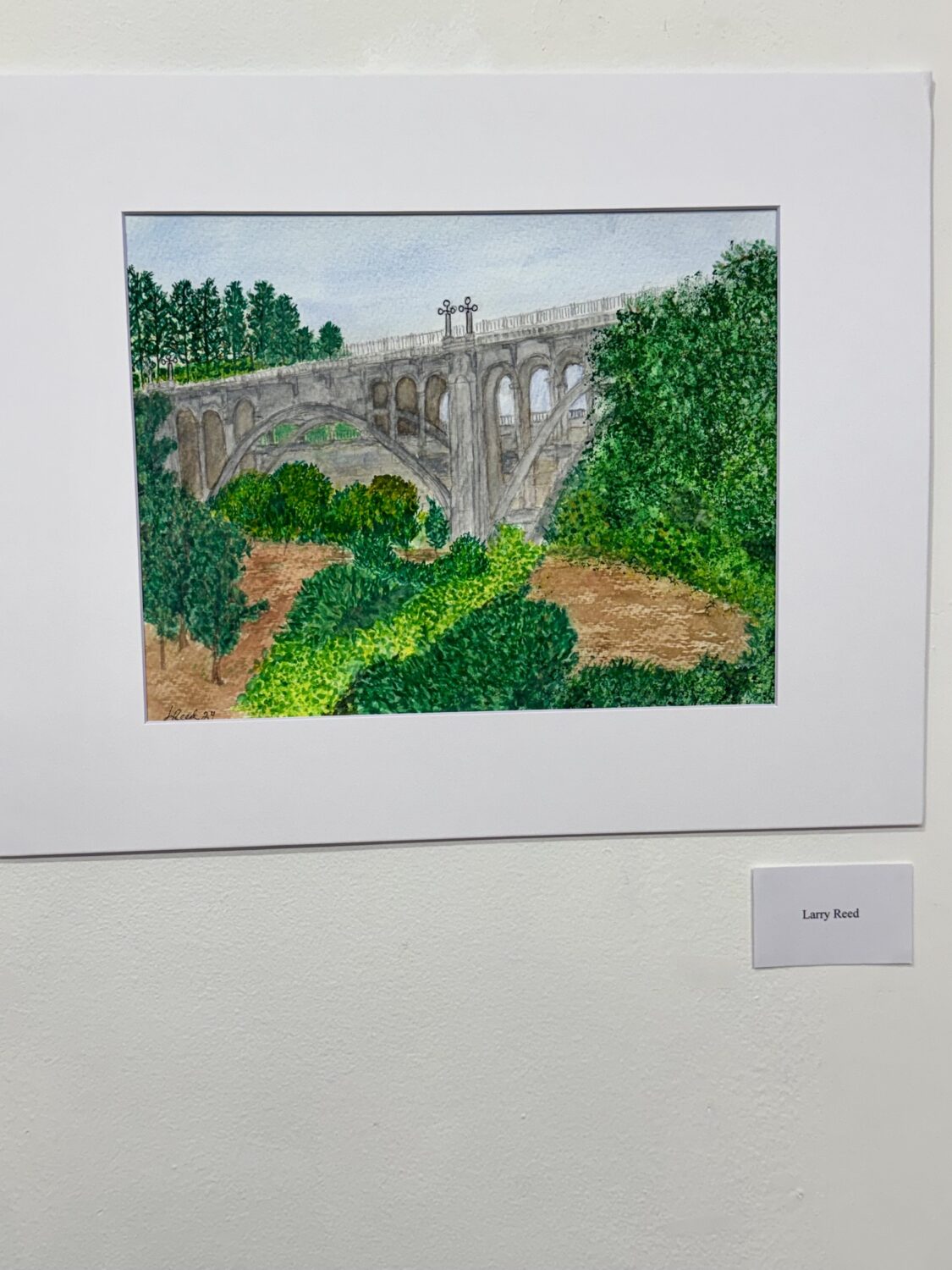 Painting of a stone arch bridge with multiple arches, set in a lush green landscape under a blue sky, framed and signed by Larry Reed.