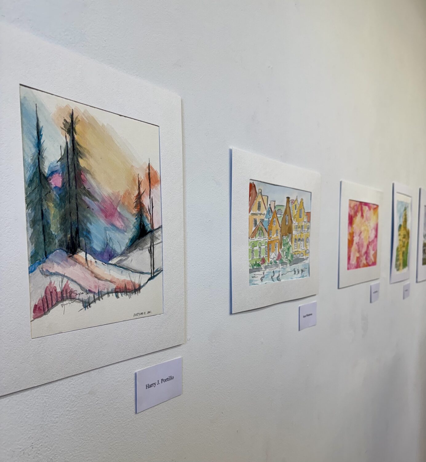 Row of colorful watercolor paintings in white frames hung along a light gallery wall, the first piece depicting a snowy landscape with tall trees and vibrant sky hues