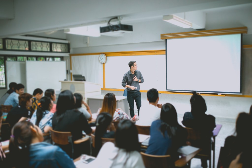 A male teacher giving a lecture to students in a university classroom with a projector screen.