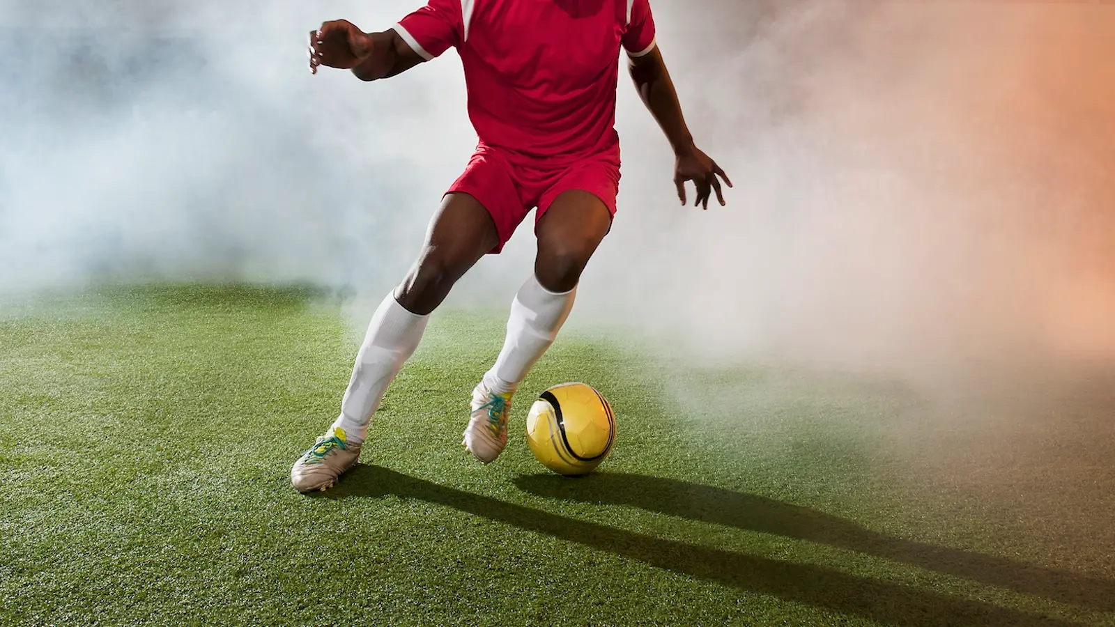 A soccer player kicks a yellow ball on turf with a haze of smoke around him