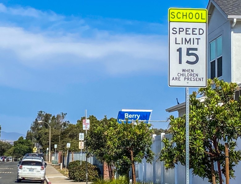 A speed limit sign on a tree-lined street shows a 15 mph limit a short distance from a school sign that reads "Berry Elementary School."