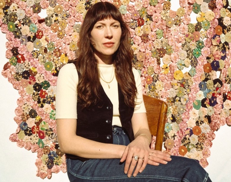 A woman in a t-shirt and vest sits with her legs crossed before a embroidered wall hanging.