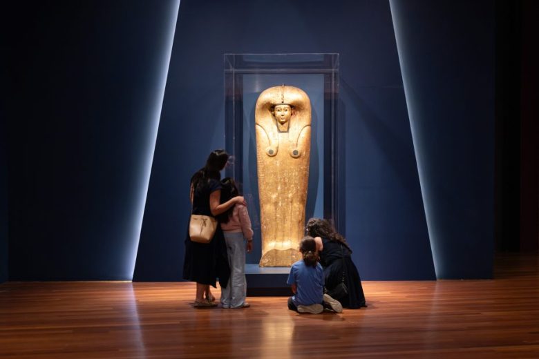 Four people look at an ancient Egyptian sarcophagus displayed upright in a glass case in a dimly lit museum gallery.