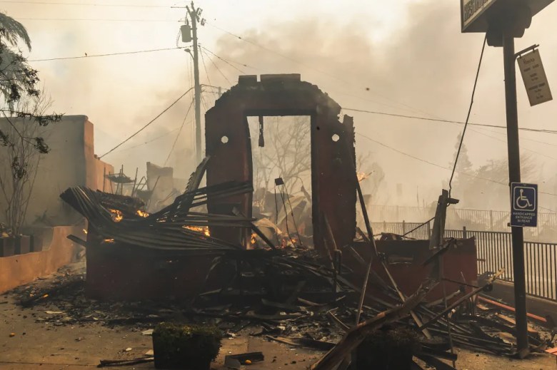 The remaining structure of a dark red building burned and destroyed by a wildfire stand amidst ruins as smoke fills the sky.