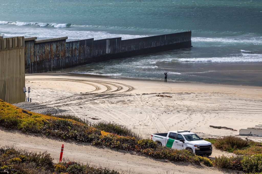 A border wall extends from the beach into the ocean as waves break along the shoreline. A U.S. Border Patrol truck is parked on a dirt road overlooking the beach, and a lone person stands near the water beside the fence.
