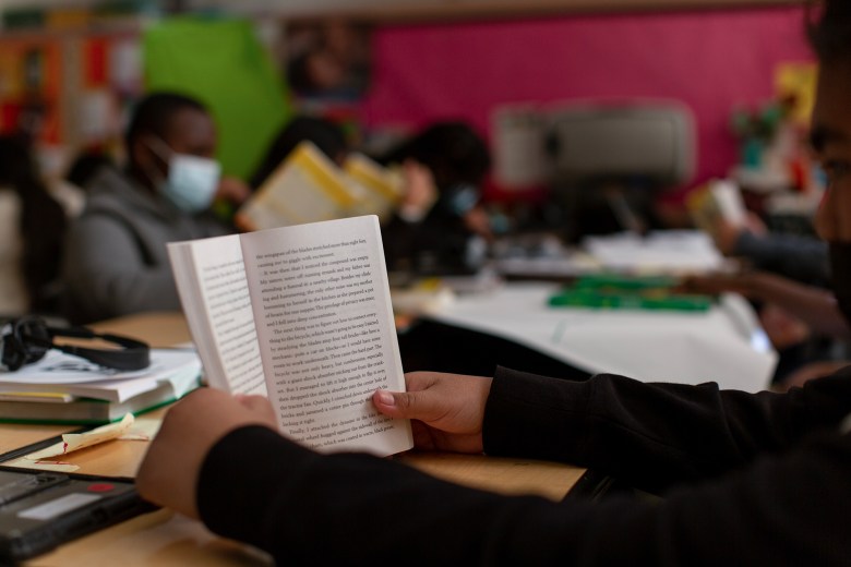 A close-up of a student holding an open book at a classroom desk, with other students reading in the background. Some students wear face masks, and the classroom walls are decorated in bright colors. Books and school supplies are scattered across the desks.