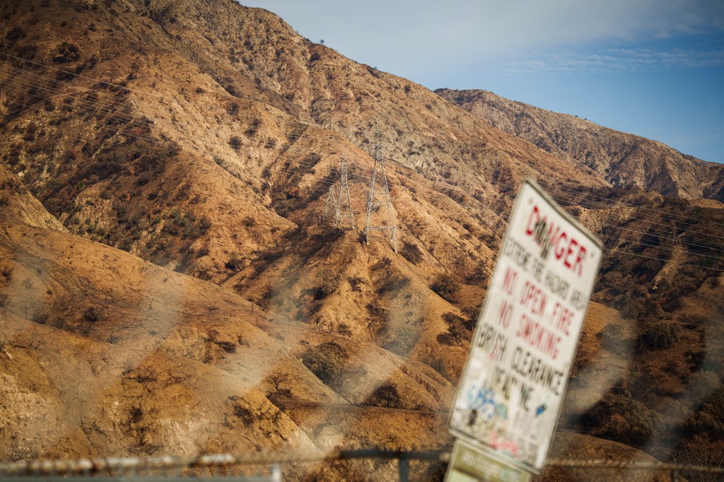 A landscape of scorched hills shows remnants of a wildfire, with dry, barren slopes dotted with blackened vegetation. Power lines and transmission towers stretch across the terrain. In the foreground, a blurred warning sign with red lettering reads "DANGER" and cautions against open fires and smoking. The hazy fence adds a sense of distance, emphasizing the vast, rugged expanse of the burned landscape under a clear sky.