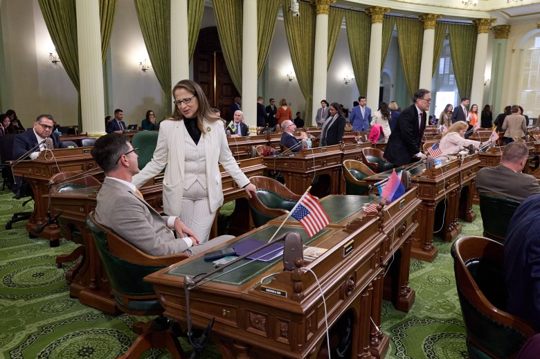 A wide-view of lawmakers standing and talking to each other in the middle of a legislative floor filled with desks. A white chandeliers hangs above the room and the room is decorated with beige and green fixtures.