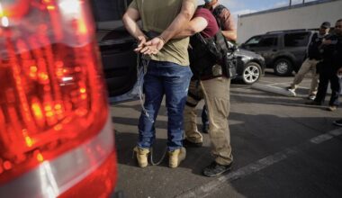 A person in jeans and a T-shirt stands while someone with a vest and a gun ties something around them.