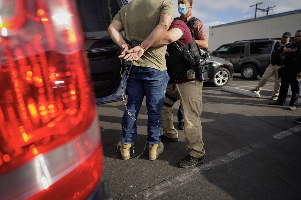 A person in jeans and a T-shirt stands while someone with a vest and a gun ties something around them.