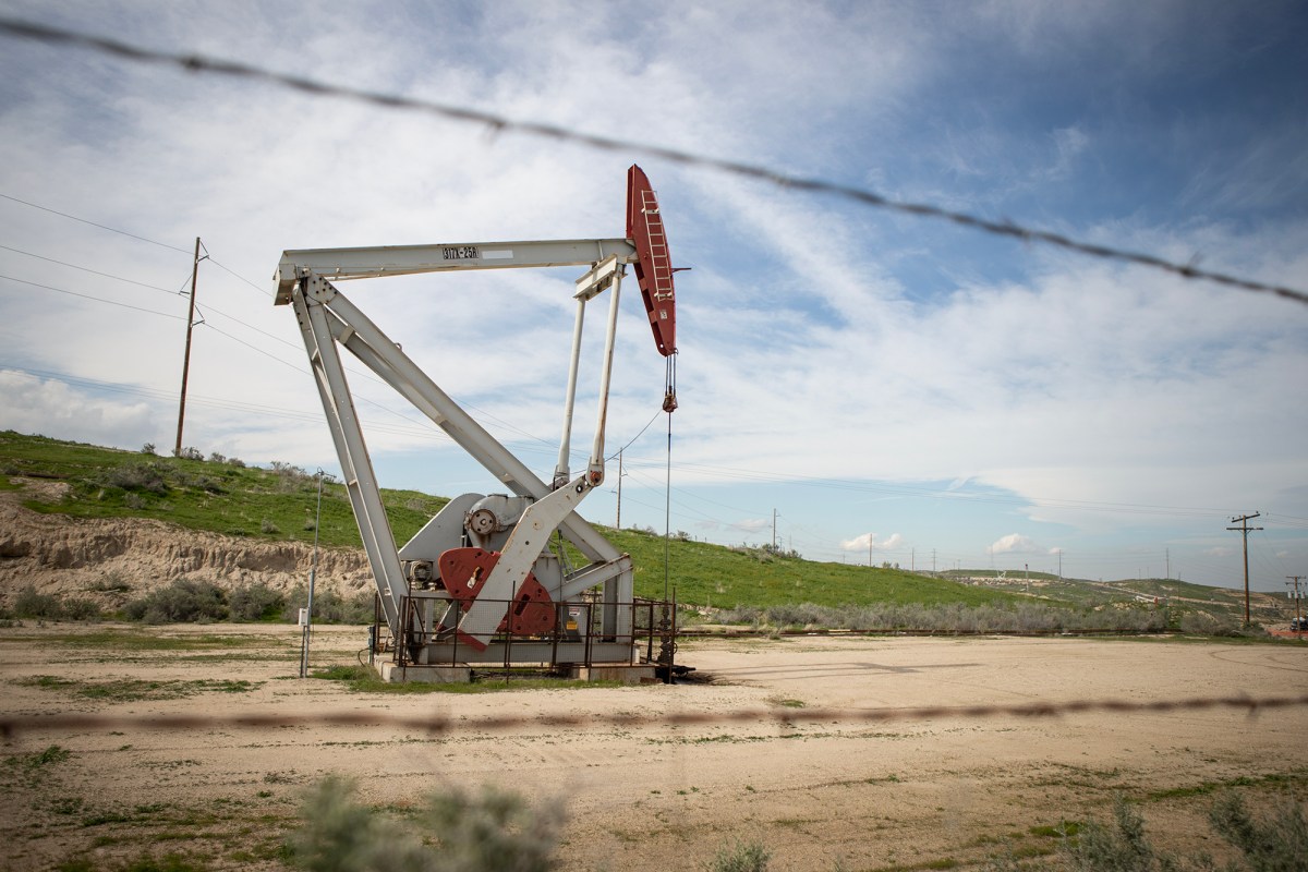 A pumpjack stands in an open oil field behind strands of barbed wire, with grassy hills, utility poles and a partly cloudy sky in the background. The large white and red oil pump sits alone on a dirt lot enclosed by a low fence.