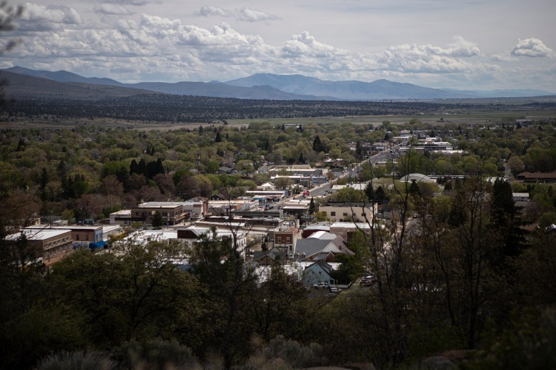 A view of downtown Susanville from a hillside surrounded by green trees and forestland, with mountain ranges in the distance.