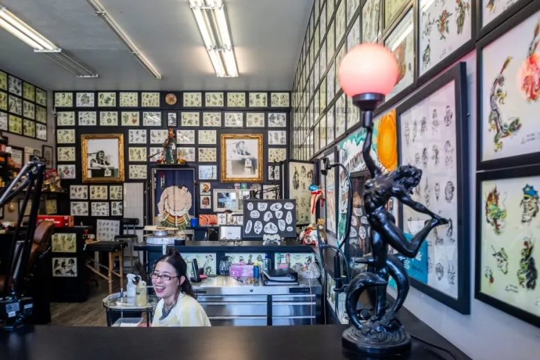 A woman sits behind the counter in a tattoo parlor, surrounded by framed tattoo designs covering the walls. A decorative lamp with a pink globe stands on the counter.