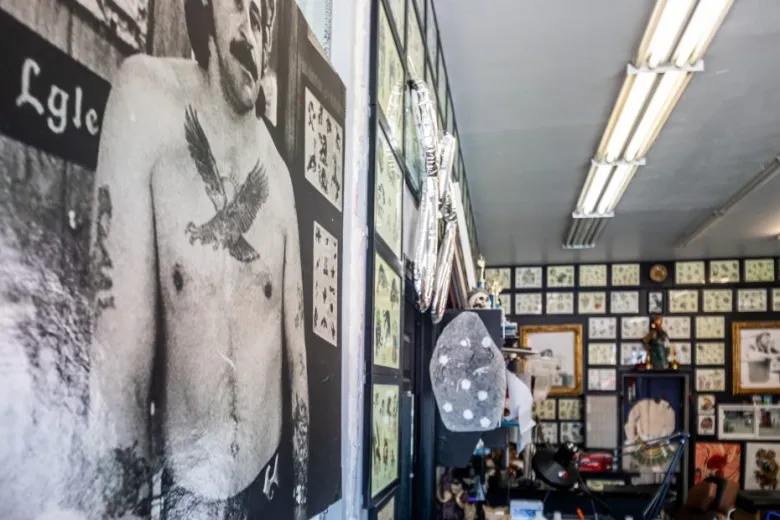 A tattoo studio interior with flash art on the walls, fluorescent lights overhead, and a large black-and-white photo of a tattooed man displayed prominently.