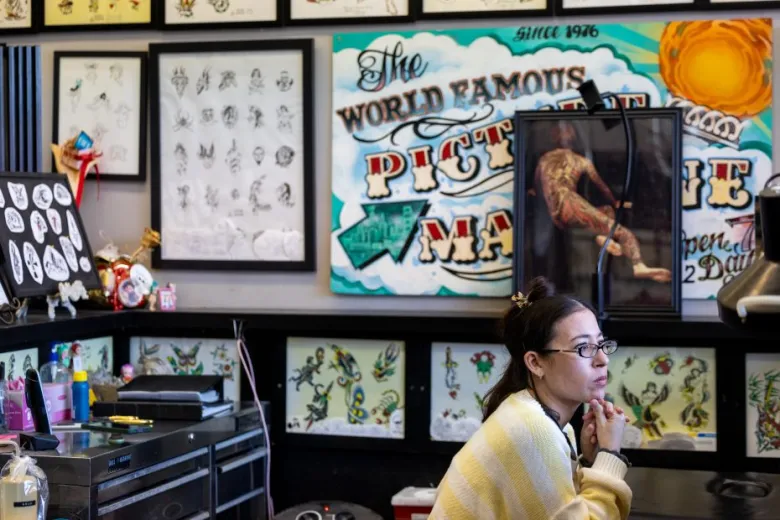 A woman sits in a tattoo studio, surrounded by tattoo designs and colorful artwork on the walls.
