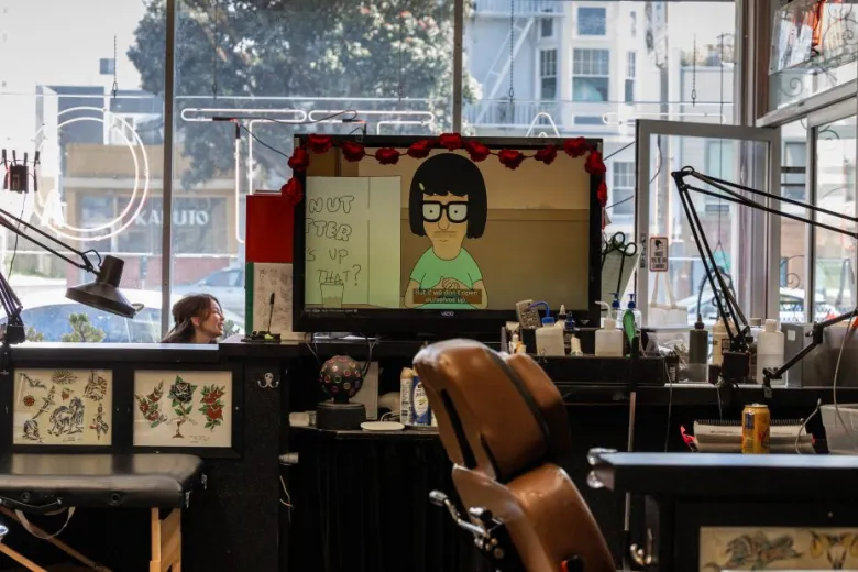 A tattoo studio interior with empty chairs, tattoo equipment, and a TV showing a cartoon character with captions, in front of a window facing the street.