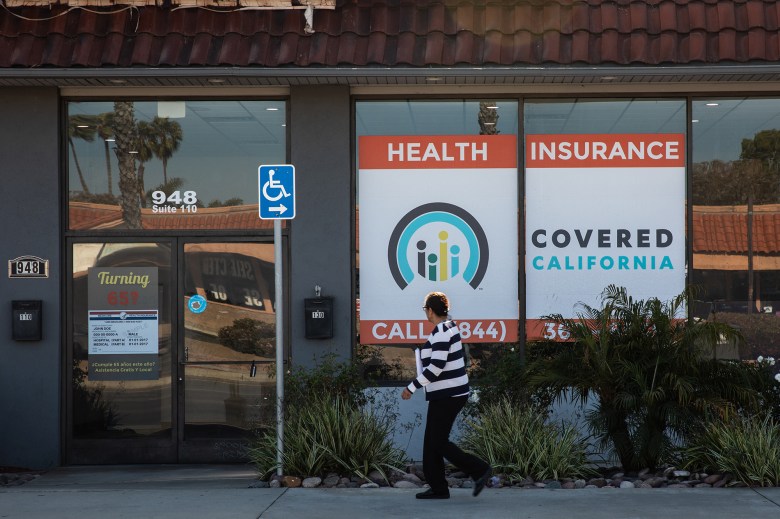 A person walks on the sidewalk in front of a red, white, dark blue, and light blue Covered California health insurance sign on a large window in Chula Vista.