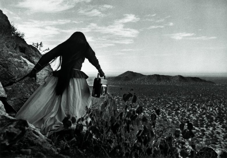 A person in a long skirt stands on rocky terrain holding a boom box, looking out over a vast desert landscape with hills in the distance.
