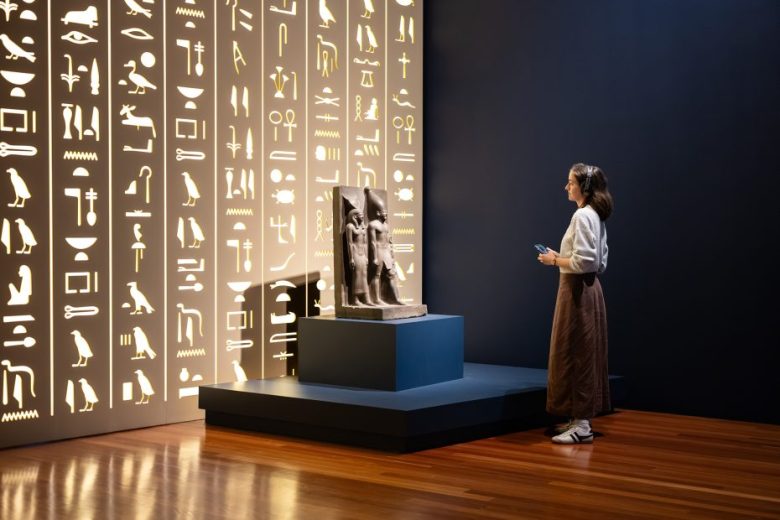A woman stands in a museum, observing an ancient statue displayed on a pedestal beside a wall illuminated with Egyptian hieroglyphs.
