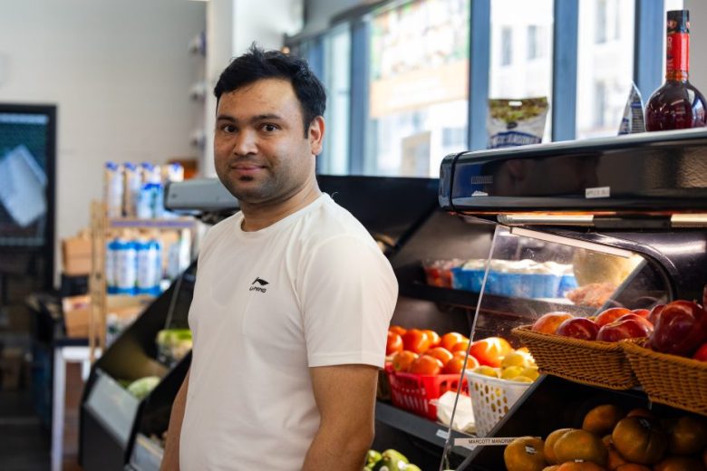 A man in a white shirt stands inside a grocery store near displays of fresh produce, including tomatoes and apples.