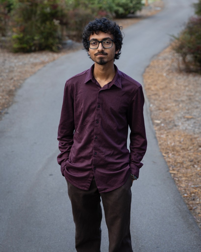 A person with curly hair and glasses stands on a paved path with their hands in their pockets, looking toward the camera. Trees and shrubs line the winding path in the softly blurred background.
