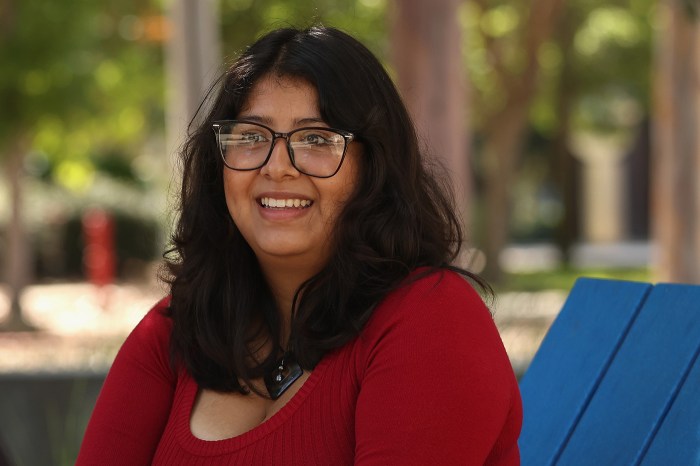 A high school student wearing glasses and a red top smiles at the camera, seated outdoors near a blue chair.