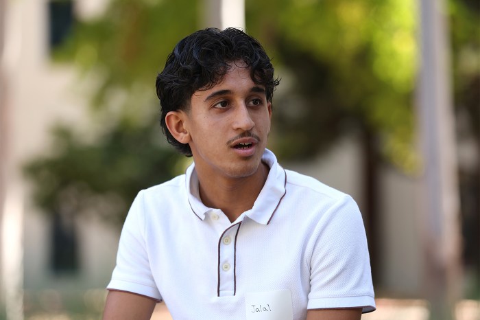 A high school student wearing a name tag and white polo shirt speaks, looking off to the side, outdoors.