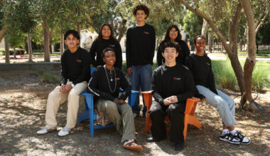 Seven high school students in matching black Northeastern sweatshirts pose together outdoors under trees, two seated in colorful chairs and the rest standing or kneeling around them.