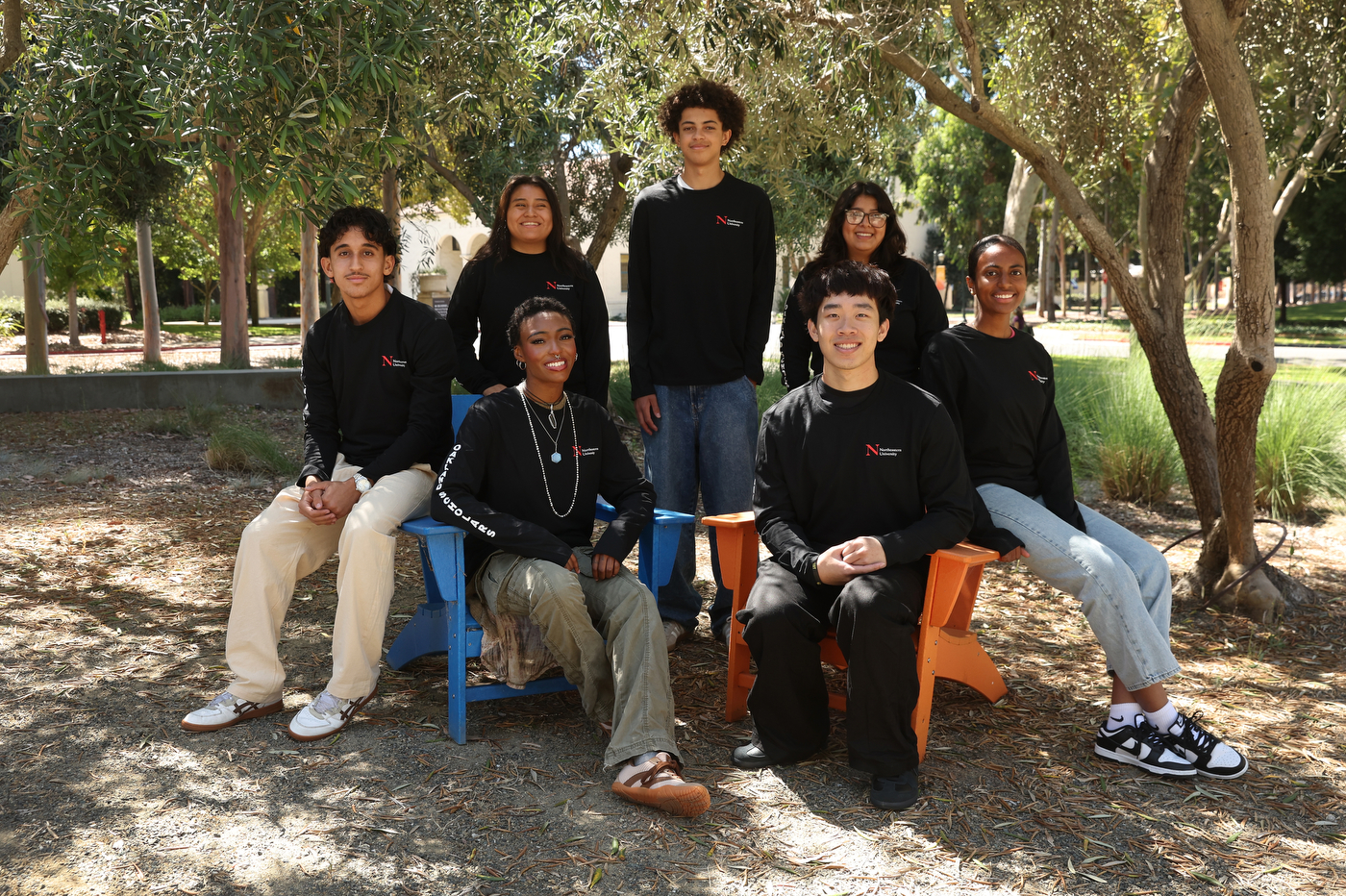Seven high school students in matching black Northeastern sweatshirts pose together outdoors under trees, two seated in colorful chairs and the rest standing or kneeling around them.