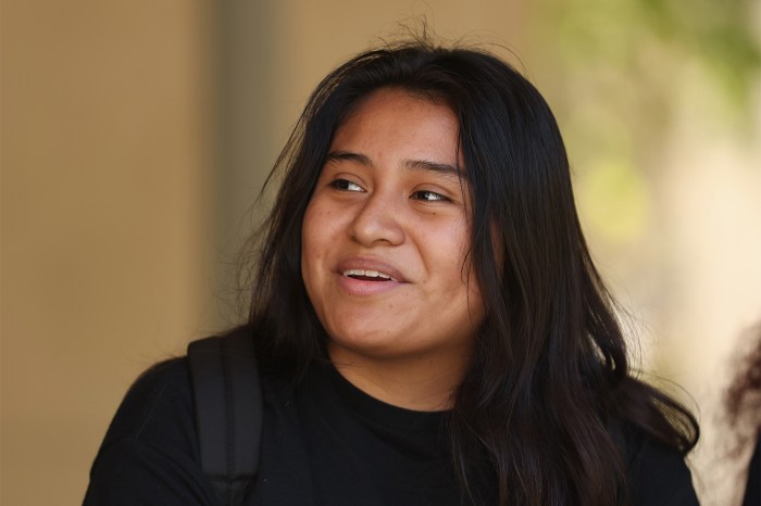 A high school student in a black sweatshirt smiles while looking off to the side, outdoors.