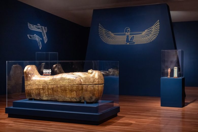 A golden Egyptian sarcophagus and several artifacts are displayed in a museum room with blue walls decorated with gold winged motifs.